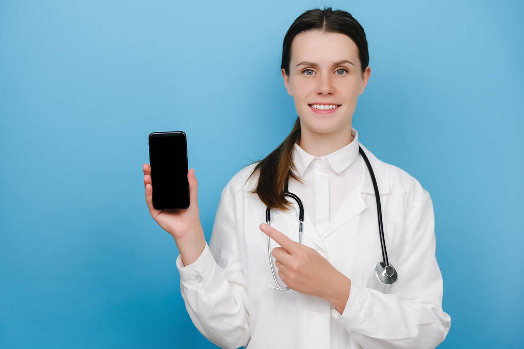 Счастливая молодая женщина-врач, указывая пальцем на мобильный экран, рекомендует скачать чикап, консультационное приложение, изолированное на синем фоне. Covid-19, медицинские работники и онлайновая медицина - Фото, изображение