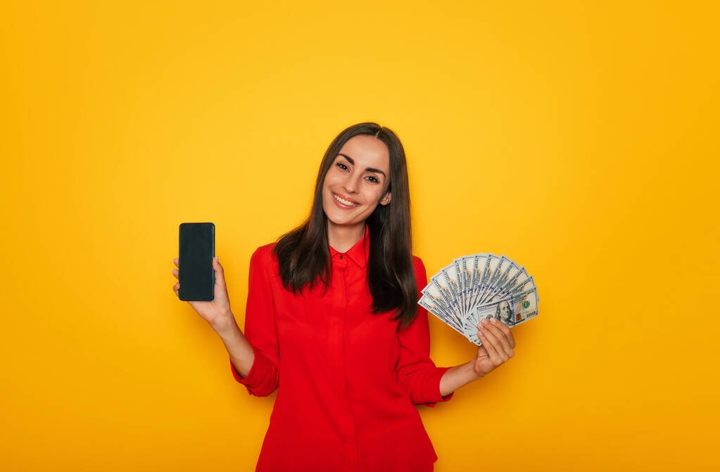 Attrayant excité sourire brune tient tas d'argent et téléphone intelligent dans les mains tout en posant sur fond jaune - Photo, image