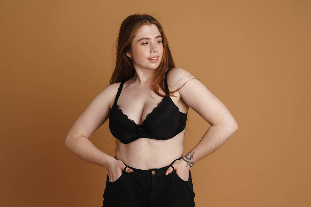 Smiling young plus size woman wearing bra posing over beige wall background - Photo, Image