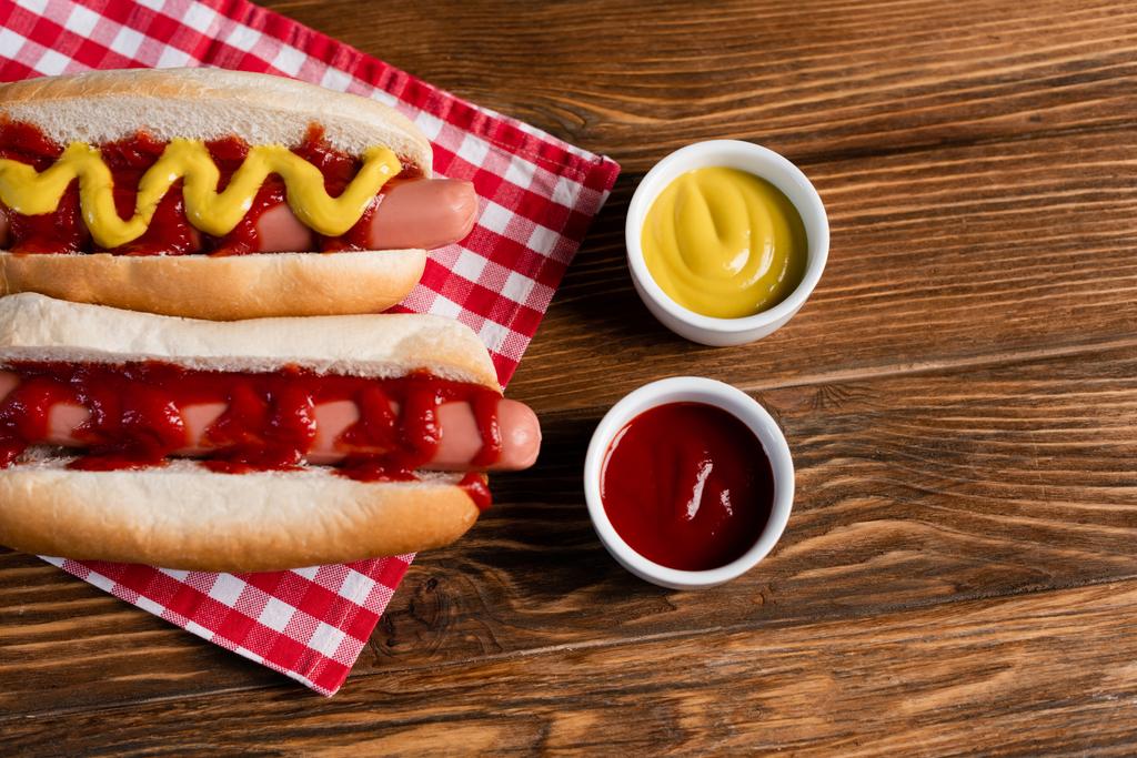 bovenaanzicht van hotdogs bij saus kommen en geruite servet op houten tafel - Foto, afbeelding