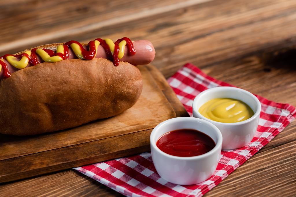 saboroso cachorro-quente em tábua de corte perto de tigelas com mostarda e ketchup e guardanapo xadrez em mesa de madeira - Foto, Imagem