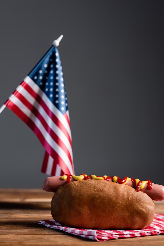 bandera americana borrosa, perrito caliente y servilleta a cuadros en mesa de madera aislada en gris - Foto, imagen