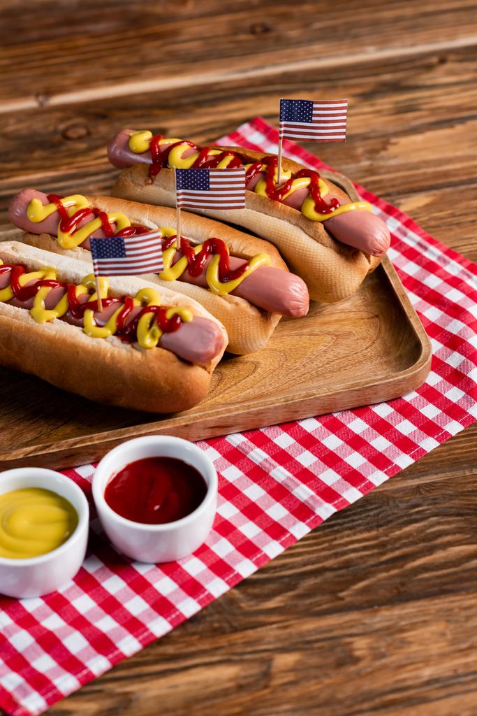 cachorros-quentes com pequenas bandeiras dos eua na bandeja e guardanapo xadrez perto de ketchup e mostarda na mesa de madeira - Foto, Imagem