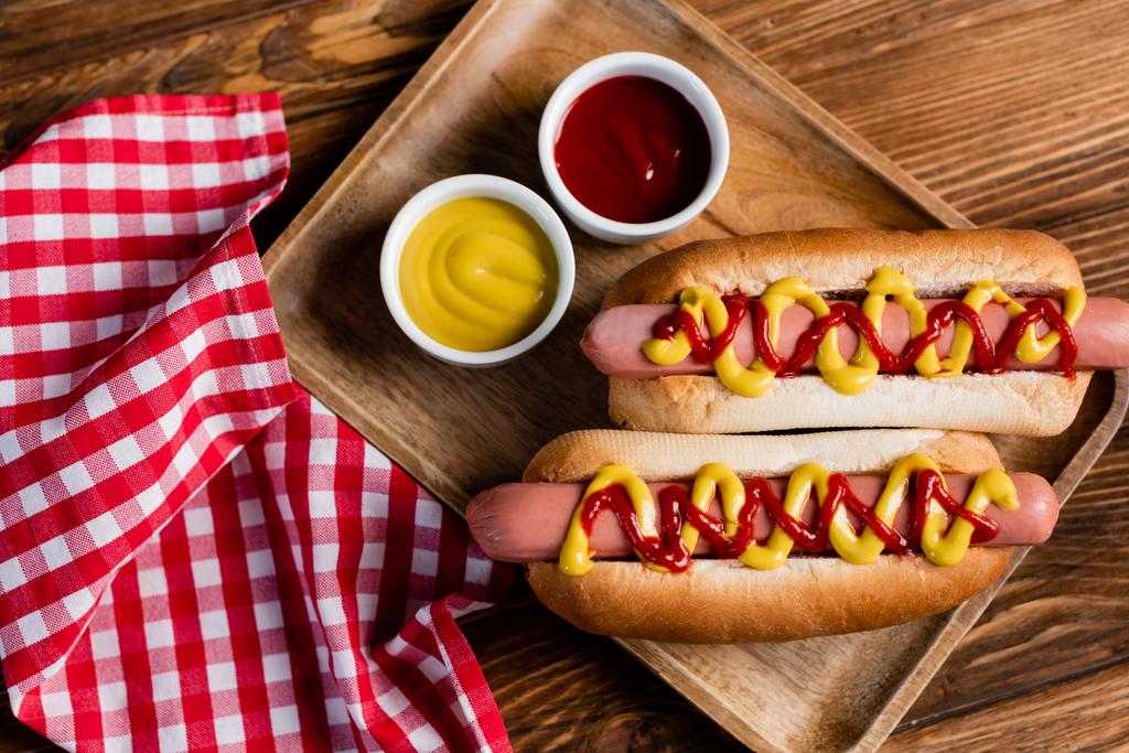 πάνω άποψη του hot dogs, κέτσαπ και μουστάρδα κοντά σε χαρτοπετσέτα καρό σε ξύλινο τραπέζι - Φωτογραφία, εικόνα