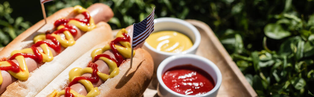 hot dogi z małą flagą usa w pobliżu miski z ketchupem i musztardą na zielonym trawniku, sztandar - Zdjęcie, obraz