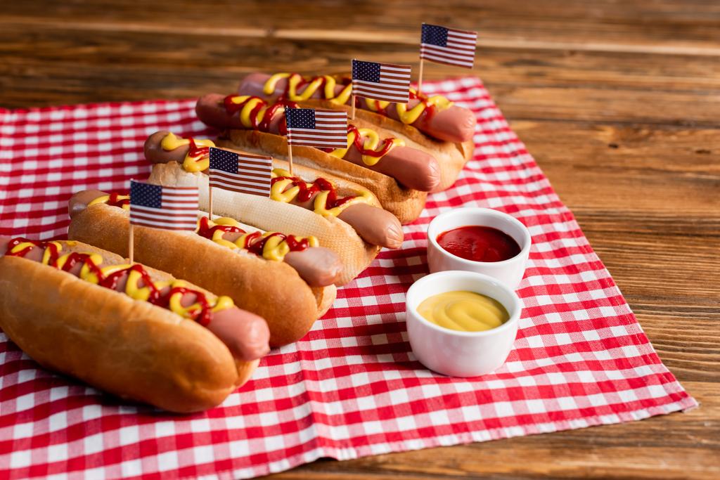 tasty hot dogs with small american flags near sauces and checkered napkin on wooden table - Photo, Image