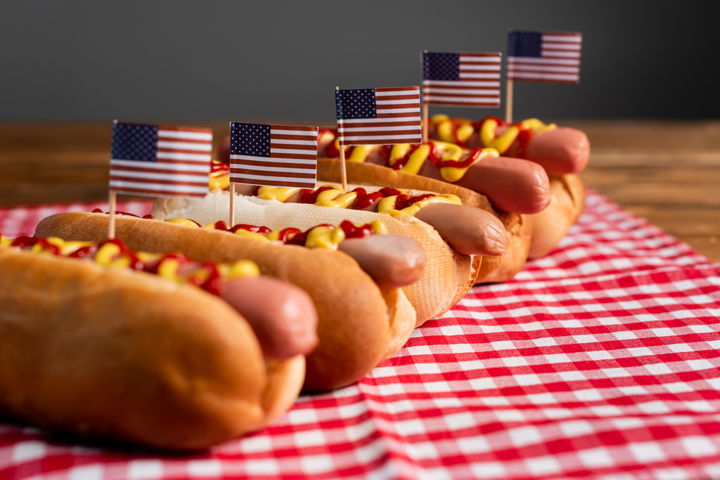 saborosos cachorros-quentes com molhos e pequenas bandeiras americanas em guardanapo de mesa xadrez isolado em cinza - Foto, Imagem