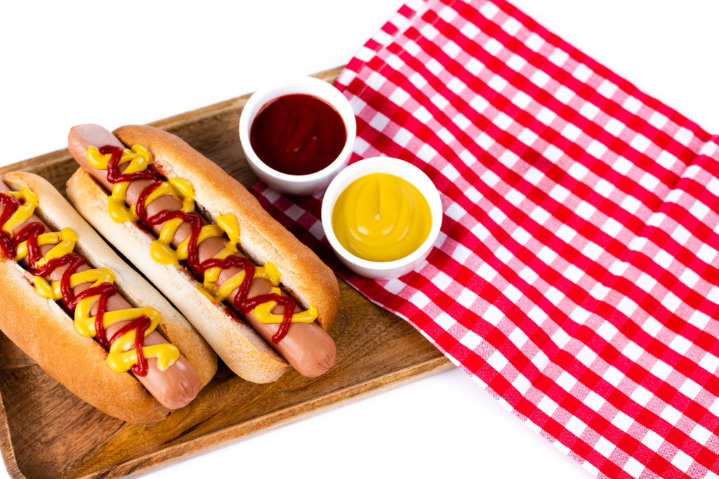 plateau en bois avec hot dogs, moutarde et ketchup près de serviette de table plaid isolé sur blanc - Photo, image