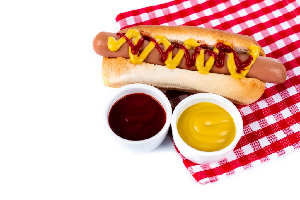miski z musztardą i ketchupem w pobliżu smacznego hot doga na serwetce w kratę na białym stole - Zdjęcie, obraz