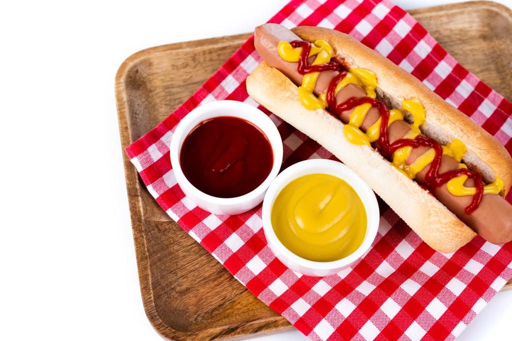 saboroso cachorro-quente com mostarda e ketchup em guardanapo de mesa xadrez e bandeja de madeira isolada em branco - Foto, Imagem
