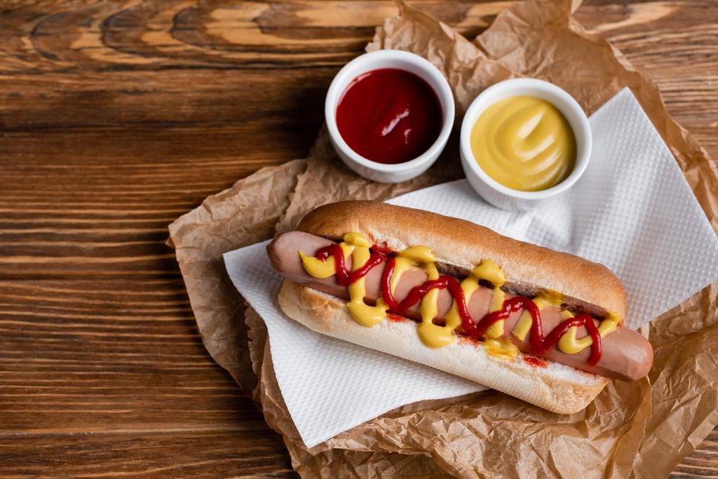 vista superior de hot dog cerca de cuencos de salsa, servilleta de papel y pergamino kraft arrugado en mesa de madera - Foto, imagen