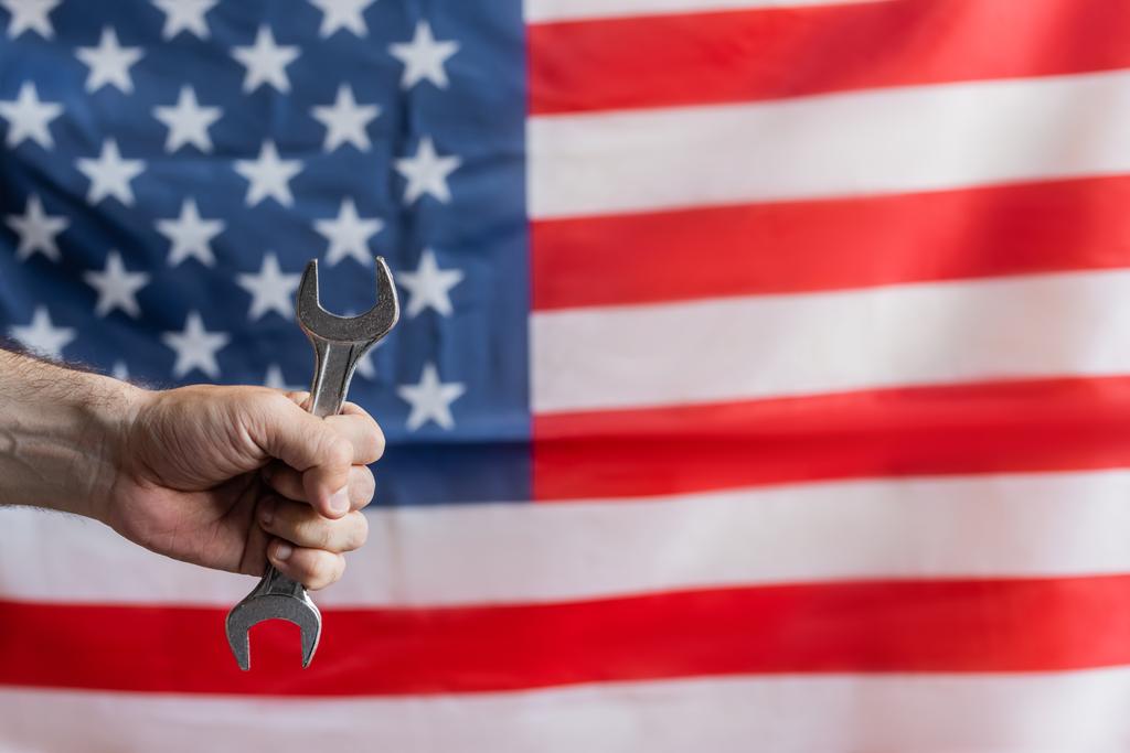 visión parcial del hombre sosteniendo la llave inglesa cerca de la bandera de EE.UU. sobre fondo borroso, concepto del día del trabajo - Foto, Imagen