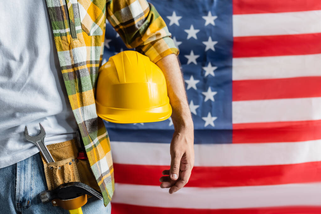 vue recadrée de l'ouvrier en chemise à carreaux et ceinture d'outils tenant un casque près du drapeau américain flou, concept de la journée du travail - Photo, image