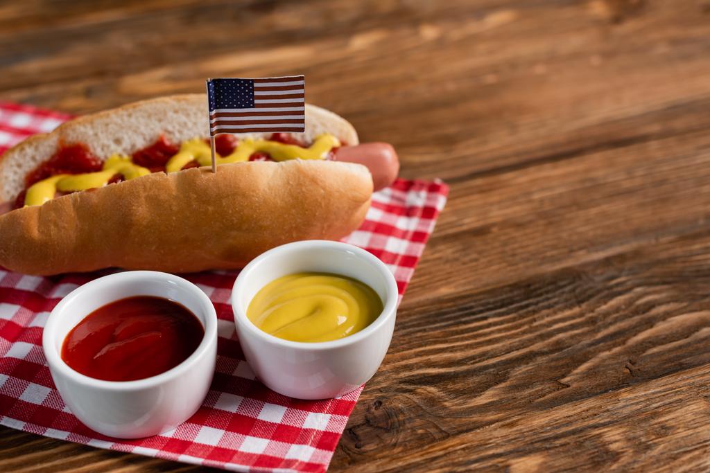 tazones con salsas cerca de sabroso perrito caliente con bandera americana pequeña y servilleta a cuadros en mesa de madera - Foto, imagen
