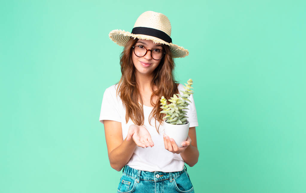 mladá hezká žena šťastně se usmívá s přátelským a nabízí a ukazuje koncept se slamákem a drží kaktus - Fotografie, Obrázek