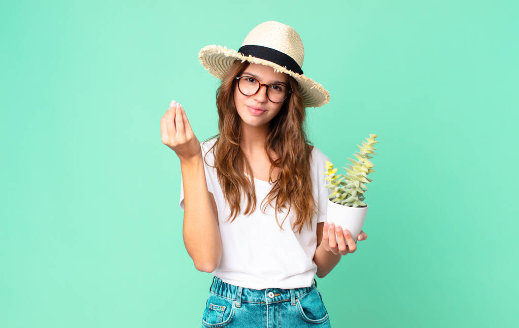 joven mujer bonita haciendo capice o gesto de dinero, diciéndole que pague con un sombrero de paja y sosteniendo un cactus - Foto, imagen