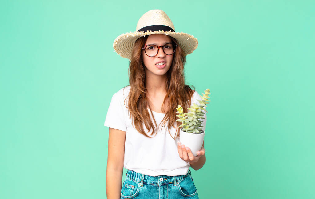 joven bonita mujer sintiéndose desconcertada y confundida con un sombrero de paja y sosteniendo un cactus - Foto, Imagen