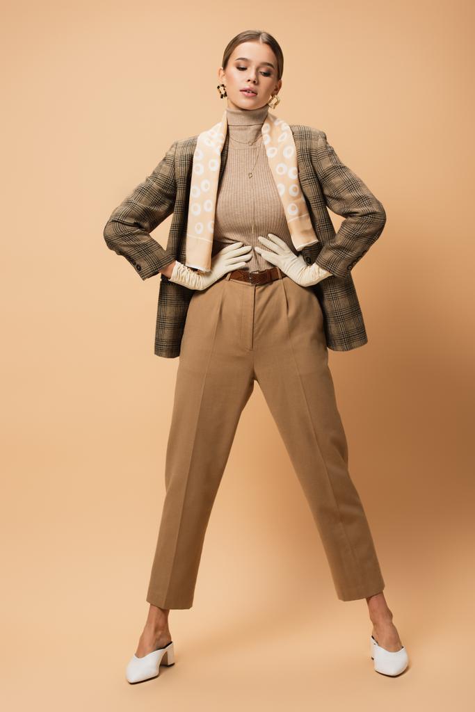 elegante mujer en chaqueta marrón y pantalones de pie con las manos en la cintura sobre fondo beige - Foto, imagen