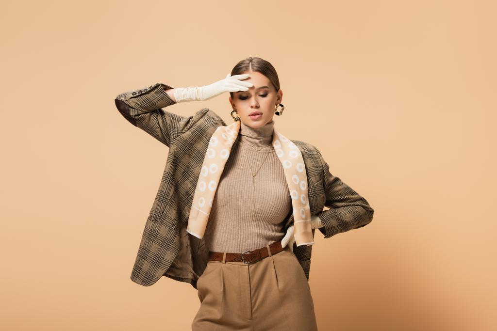 trendy woman in blazer and pants touching forehead while posing with hand on hip on beige  - Photo, Image