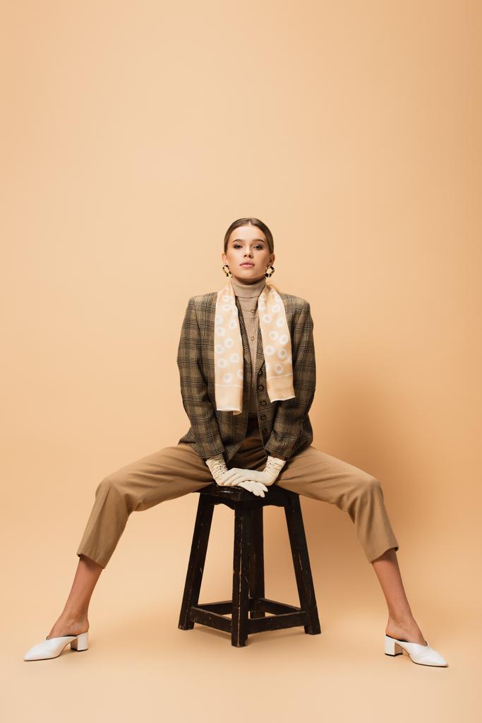 vista frontal da mulher elegante em blazer marrom e calças posando em banquinho de madeira em bege  - Foto, Imagem