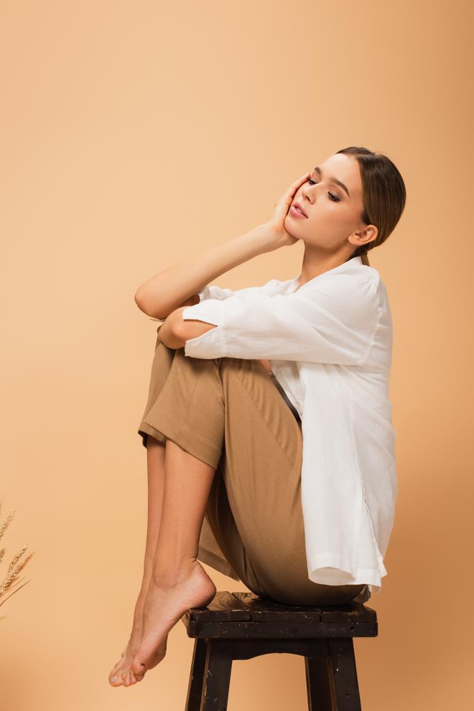 barefoot woman holding hand near face while sitting on wooden stool on beige background - Photo, Image