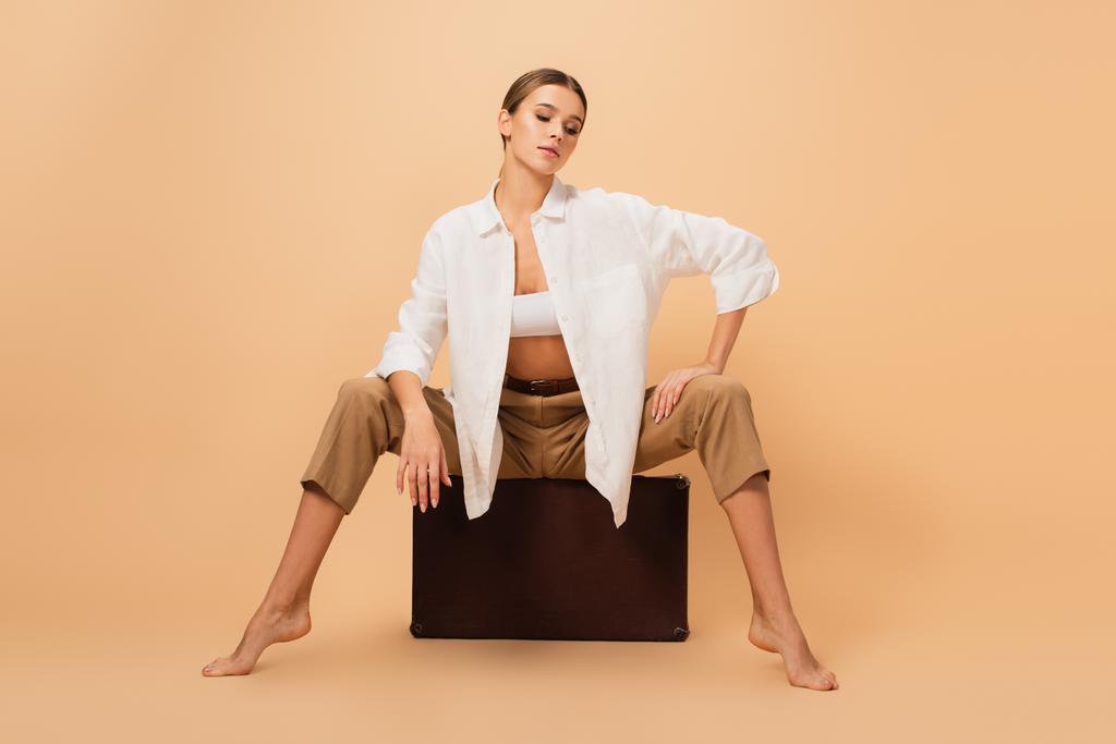 stylish barefoot woman in trousers and white shirt posing on vintage suitcase on beige background - Photo, Image