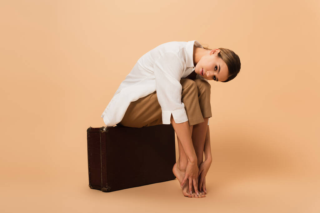 Barfüßige Frau in Hemd und Hose berührt Beine, während sie auf einem Retro-Koffer auf beigem Hintergrund sitzt - Foto, Bild