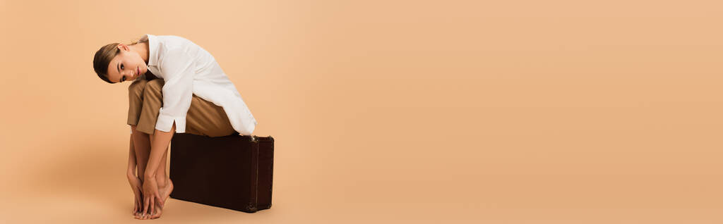mezítlábas nő elegáns ruhák ül vintage bőrönd bézs háttér, banner - Fotó, kép