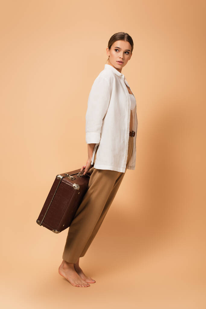 barefoot woman in pants and white shirt standing on tiptoe with vintage suitcase on beige background - Photo, Image