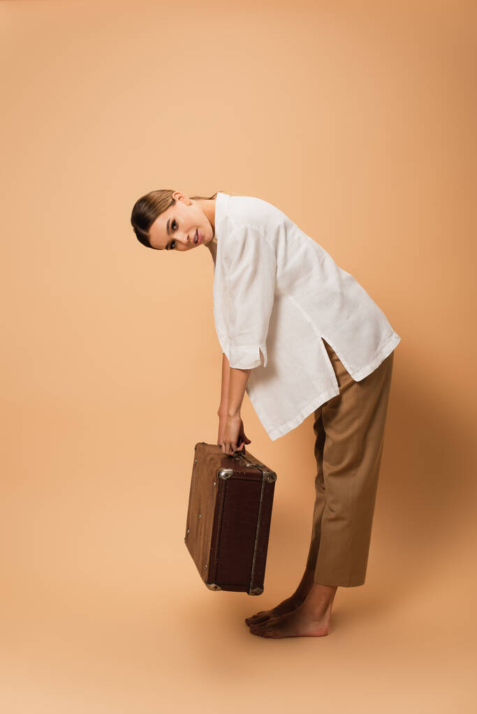 Pantolon ve beyaz gömlekli çıplak ayaklı kadın bej arka planda retro valiziyle bükülüyor. - Fotoğraf, Görsel