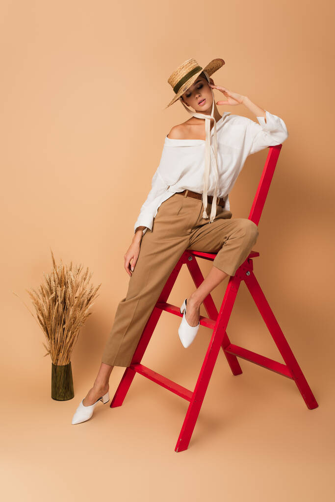 bella donna in camicia bianca, pantaloni e cappello di paglia in posa su scala rossa vicino vaso con spighette su beige - Foto, immagini