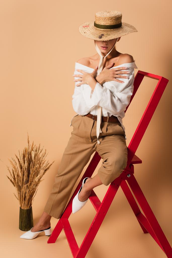 sensual woman in shirt, pants and straw hat hugging herself while sitting on red ladder on beige - Photo, Image