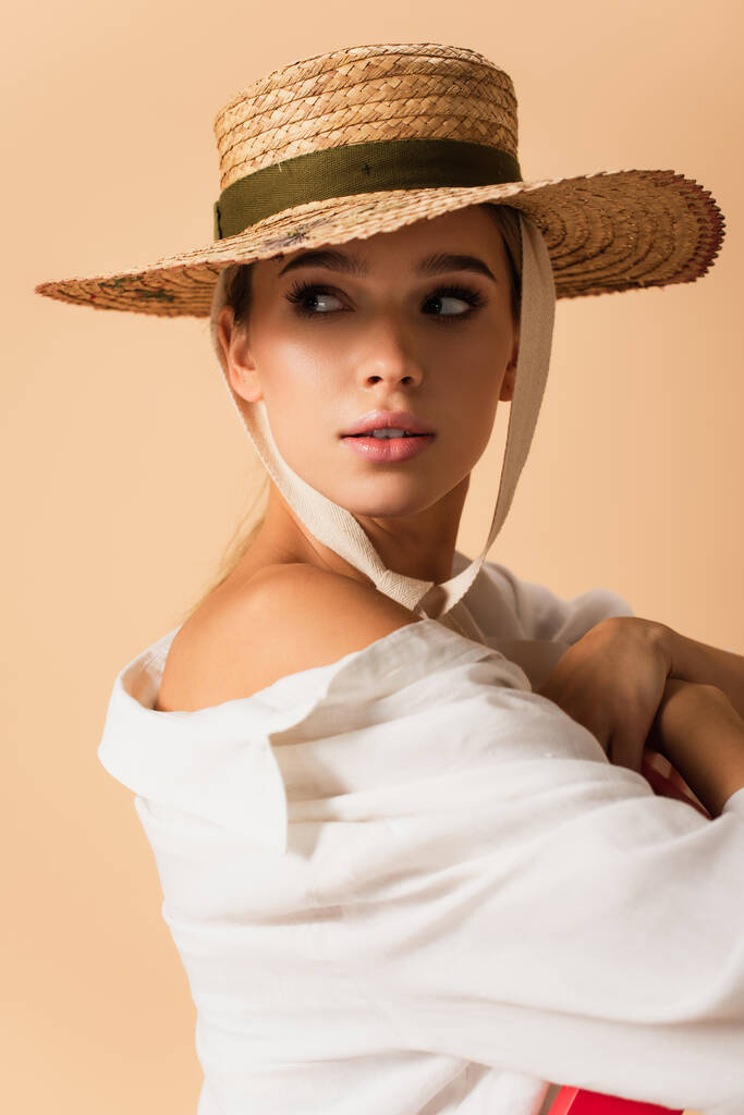 красивая женщина в белой рубашке и соломенной шляпе позирует с обнаженным плечом изолированы на бежевый - Фото, изображение