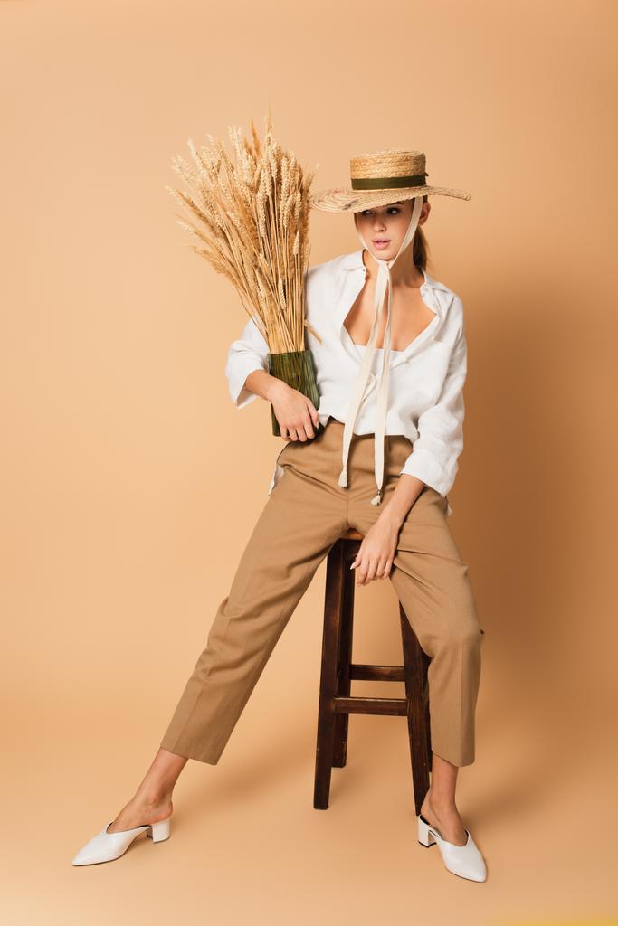 módní žena v slamáku sedí na dřevěné stoličce s vázou z hřeby na béžové  - Fotografie, Obrázek