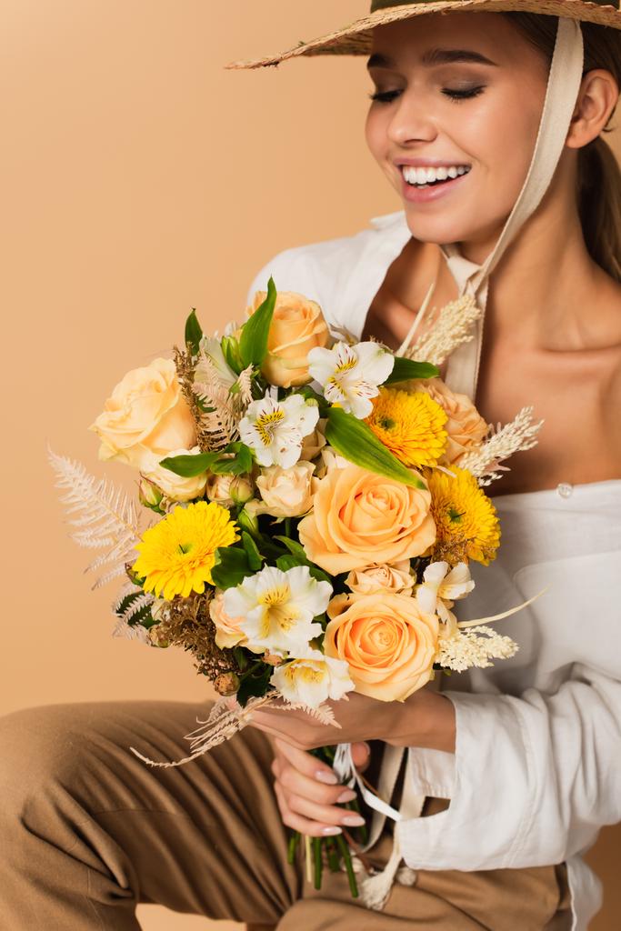 mujer feliz joven en sombrero de paja que sostiene ramo de diferentes flores aisladas en beige   - Foto, imagen