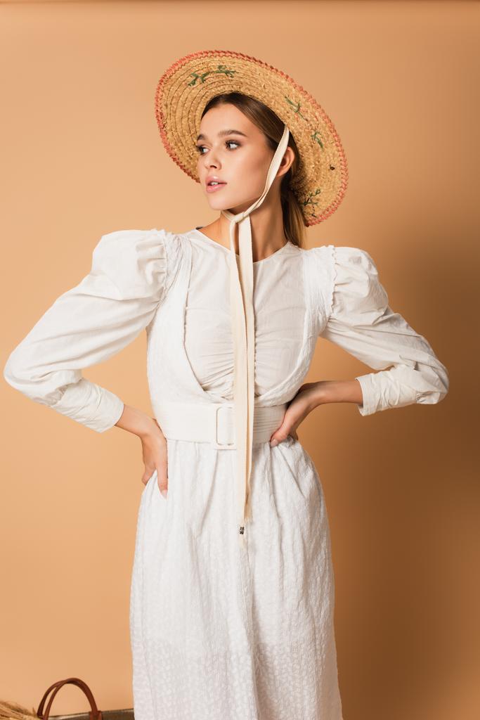 young woman in white dress and straw hat standing with hands on hips on beige  - Photo, Image