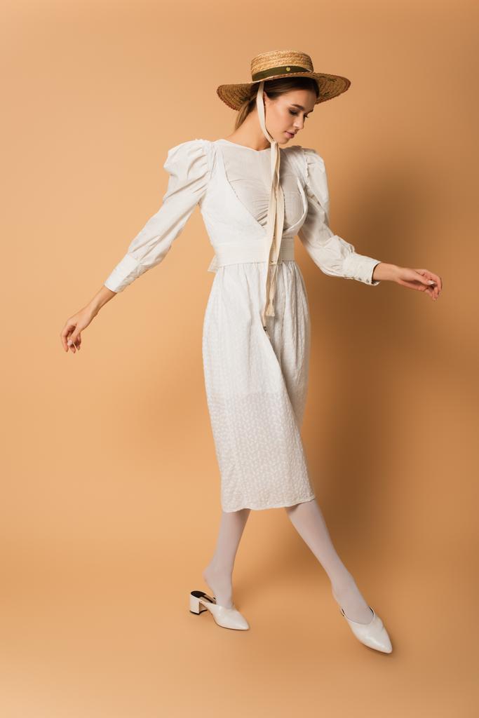 junge Frau in weißem Kleid und Strohhut posiert in voller Länge auf beige  - Foto, Bild