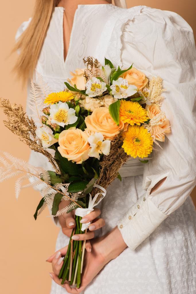 vista cortada de jovem mulher em vestido segurando buquê de flores atrás de costas em bege - Foto, Imagem