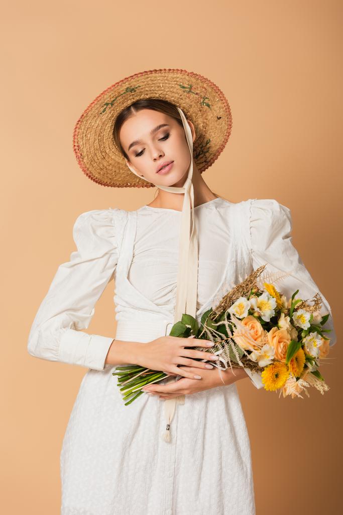 młoda kobieta w białej sukience i słomy kapelusz gospodarstwa bukiet kwiatów na beżu - Zdjęcie, obraz