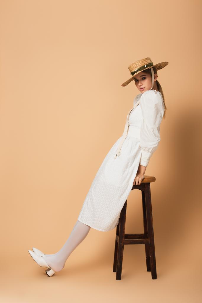 longueur pleine de jeune femme en robe blanche et chapeau de paille appuyé sur tabouret sur beige - Photo, image