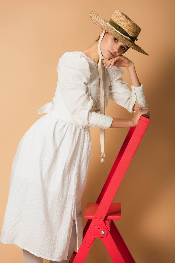 Beyaz elbiseli ve hasır şapkalı genç bir kadın bej rengi merdivenin üzerinde duruyor. - Fotoğraf, Görsel