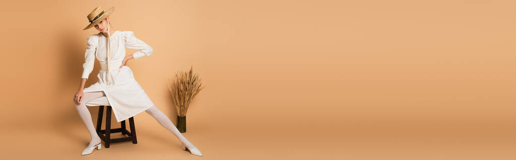 longitud completa de la mujer joven en sombrero de paja posando con la mano en la cadera cerca de la silla de madera y espiguillas de trigo en beige, bandera - Foto, Imagen