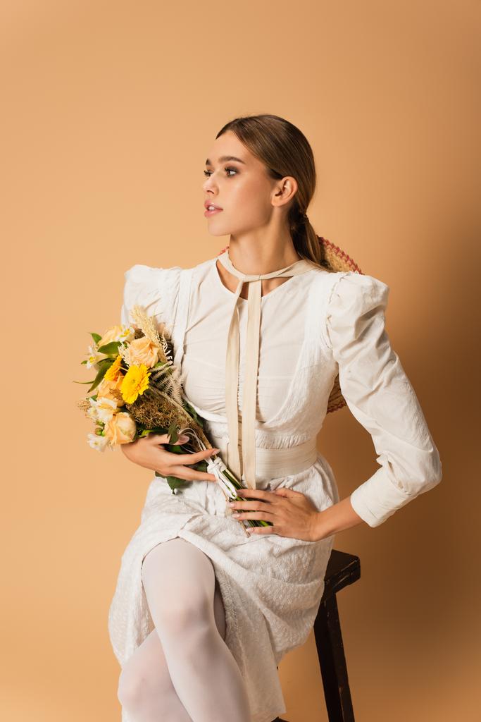 giovane donna in abito bianco che tiene bouquet con fiori e posa su beige - Foto, immagini