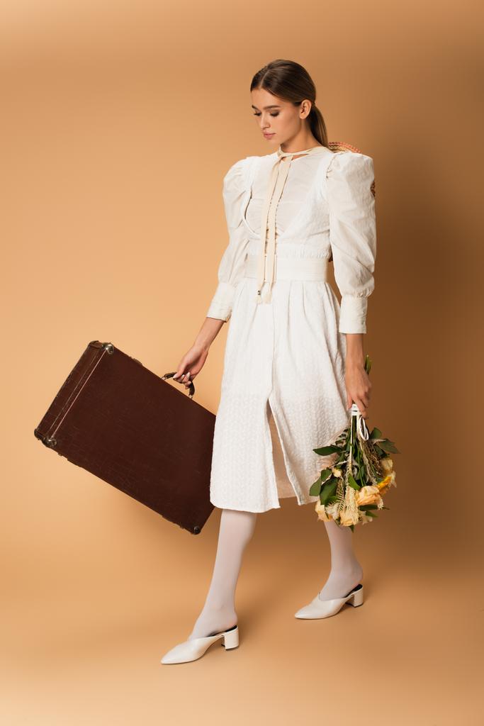 повна довжина молодої жінки в білій сукні тримає букет з квітами і валізою на бежевому
 - Фото, зображення