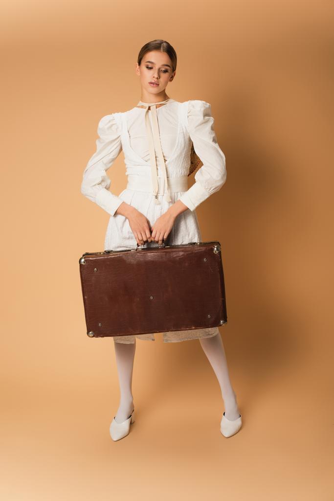 full length of young woman in white dress holding suitcase on beige - Photo, Image
