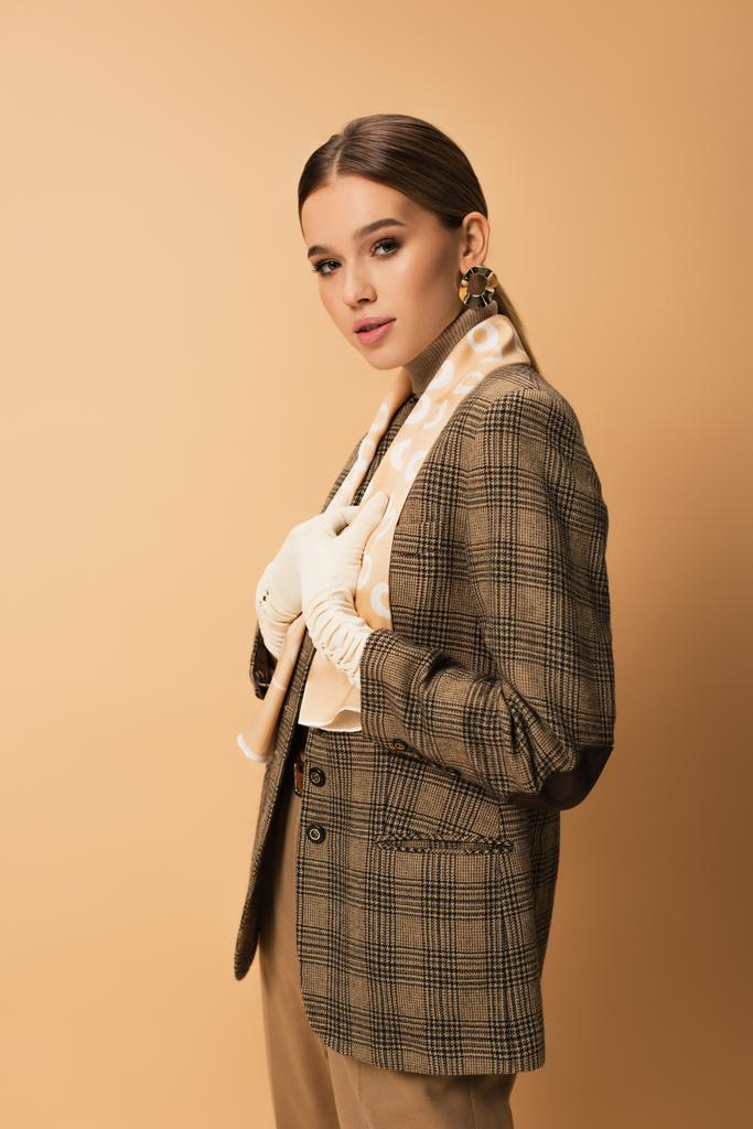 stylish woman in checkered blazer, pants and white gloves posing on beige - Photo, Image