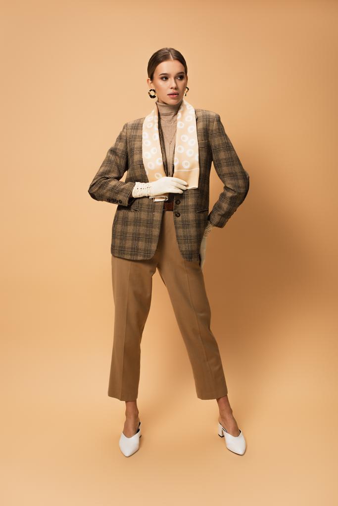 full length of young woman in checkered blazer, pants and white gloves posing on beige - Photo, Image