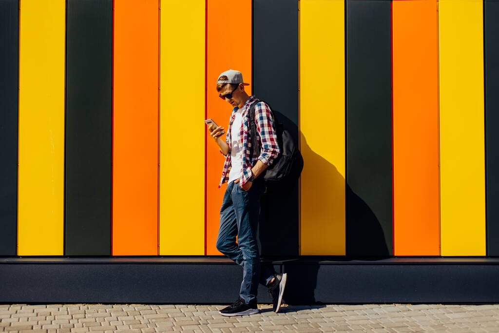 Young smiling stylish man in sunglasses and a cap, uses a mobile phone, leaning against a colorful wall, in the city - Photo, Image