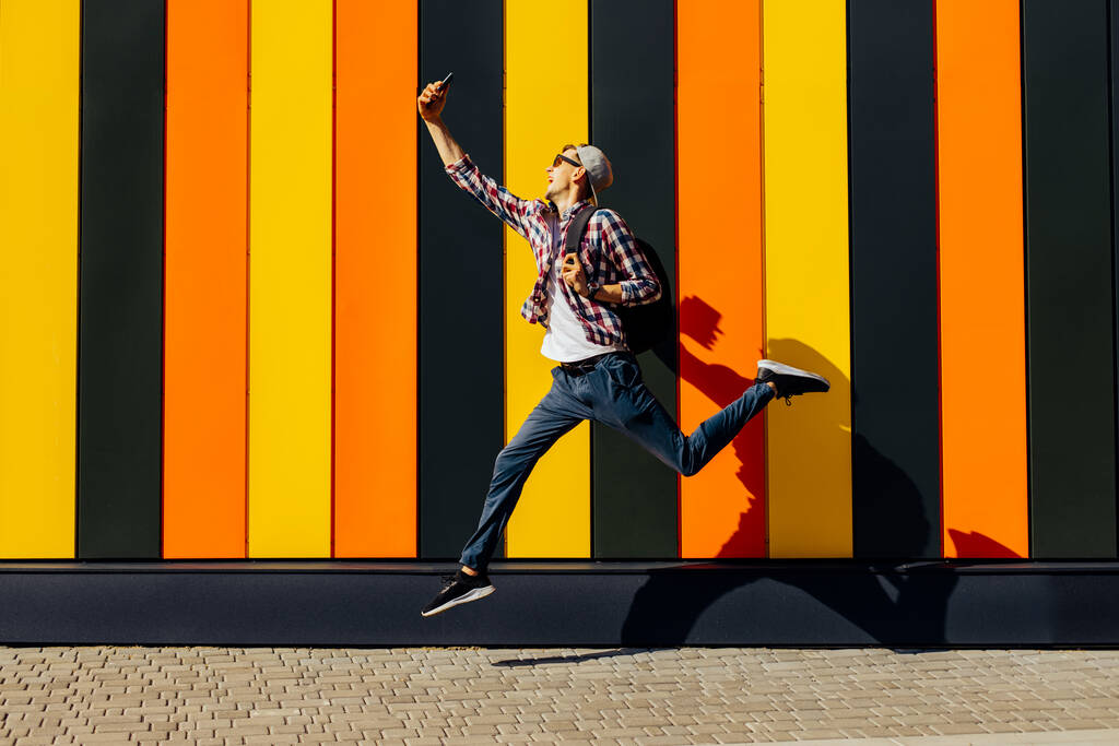 Photo of cheerful positive excited running jumping hipster with briefcase talking on mobile phone video call, isolated urban background of colorful wall - Photo, Image