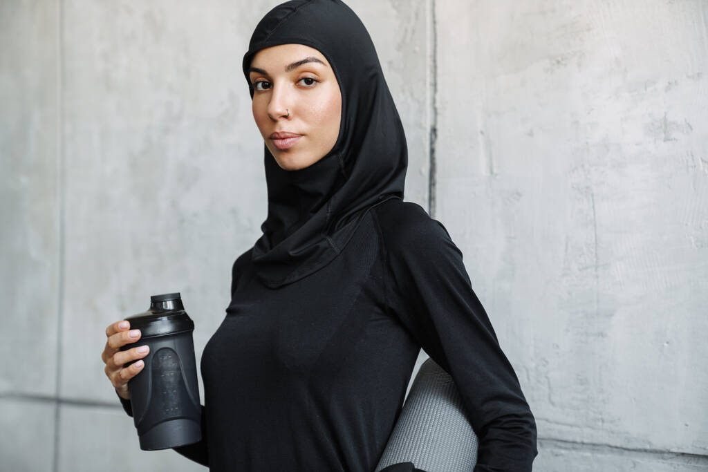 Jovem mulher muçulmana em pé hijab com tapete de fitness e garrafa de água dentro de casa - Foto, Imagem